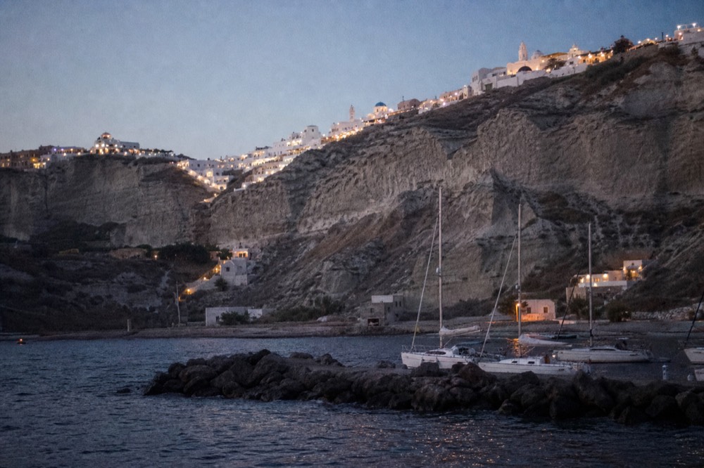 Sculpted white and grey cliff formations above a small marina breakwater, fishing boats moored in a narrow harbour under hard midday light