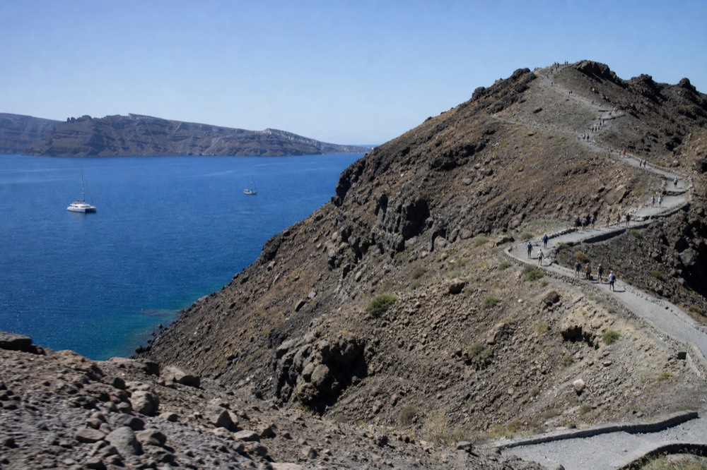 Dark volcanic crater ridge with a worn switchback path climbing it, bare grey rock and scattered walkers small on the slope