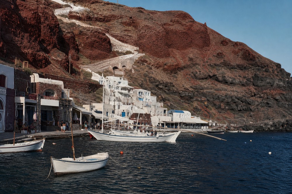 Red volcanic cliff falling to a tiny fishing harbour, whitewashed steps zigzagging up the rock face above moored wooden boats in late afternoon shade