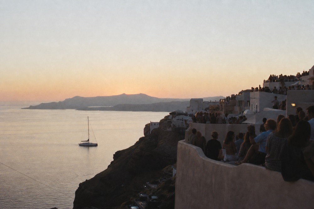 Dense silhouettes packed along whitewashed cliff-top walls of a Cycladic village at dusk, seen from a distance with warm haze over the water below