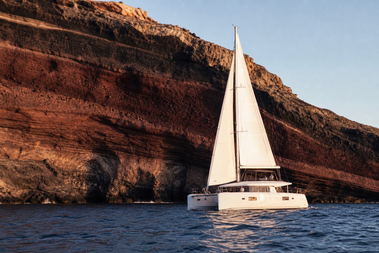White catamaran under full sail close beneath a striped volcanic cliff wall, layered red and black rock towering over dark blue water in afternoon light