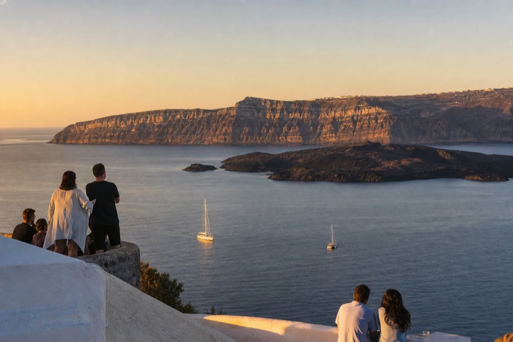 Wide caldera panorama from a rim terrace at golden hour, two dark volcanic islets sitting in still water below sheer striped cliffs
