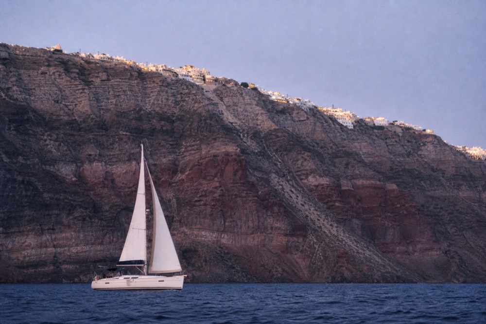 White sails hard against a towering banded caldera wall, the boat small beneath layers of red and grey volcanic rock in late light