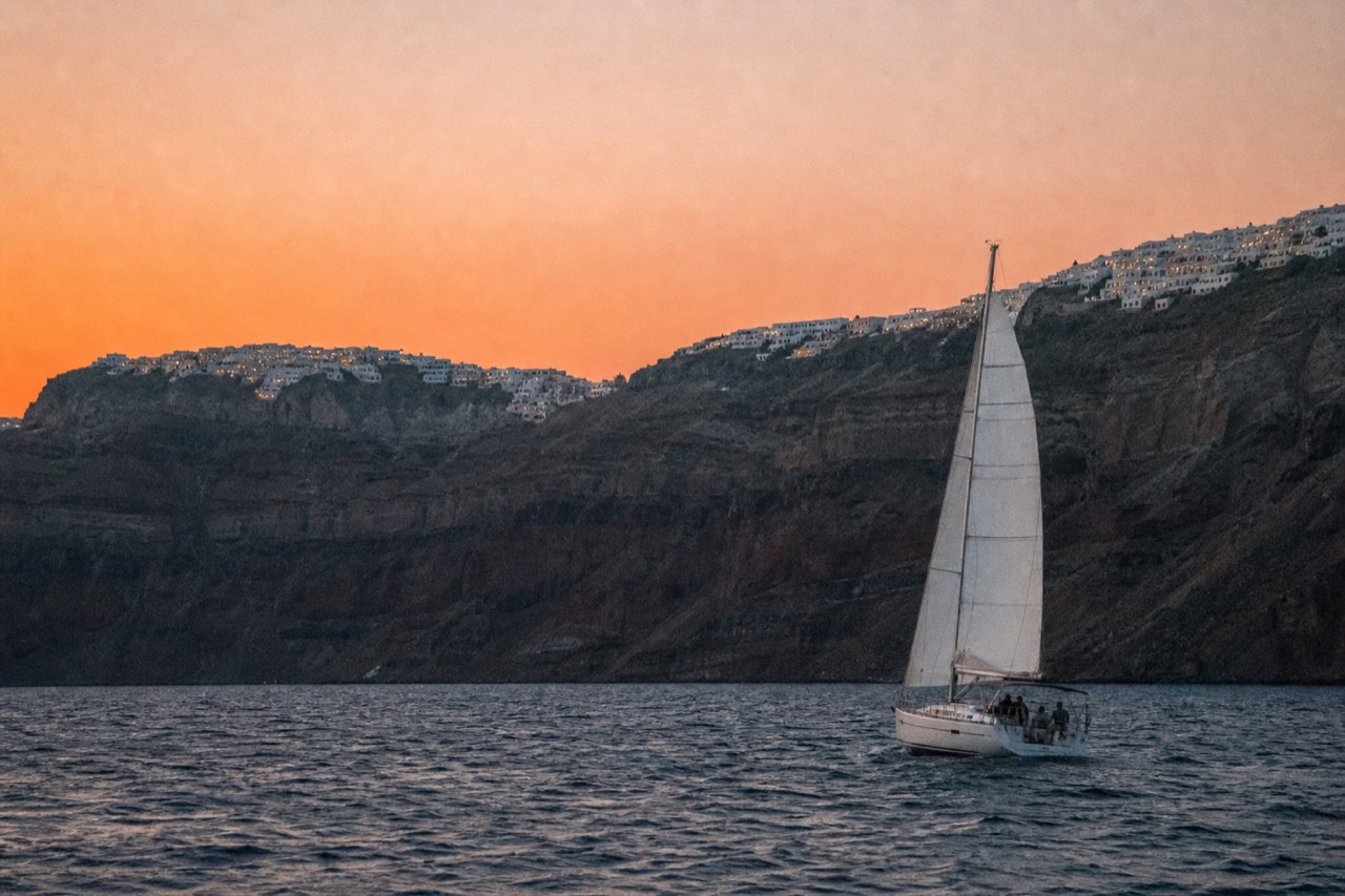 Two whitewashed clifftop villages glowing on a high volcanic rim at dusk, seen from low on the water with a band of orange light along the sea horizon