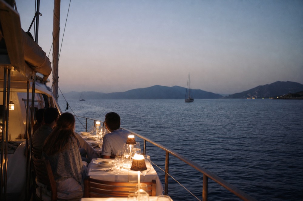 A long table set for dinner on an open deck at dusk, white cloth, glasses and small plates, warm lamps against a darkening sea