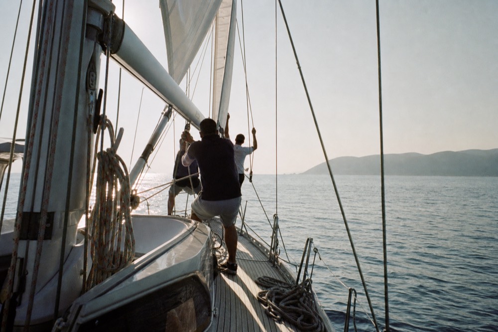 Crew hauling on a halyard as a mainsail rises in afternoon light, teak deck and coiled ropes in the foreground, calm blue water beyond
