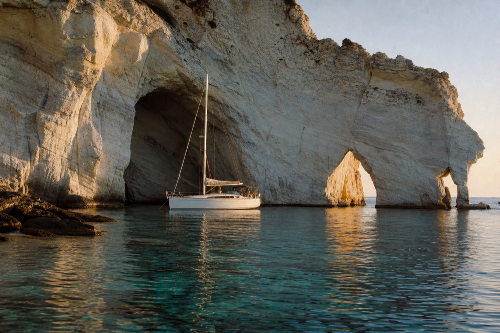 Sunlight cutting into a white limestone sea cave, a small boat drifting on glass-clear turquoise shallows beneath sculpted rock arches