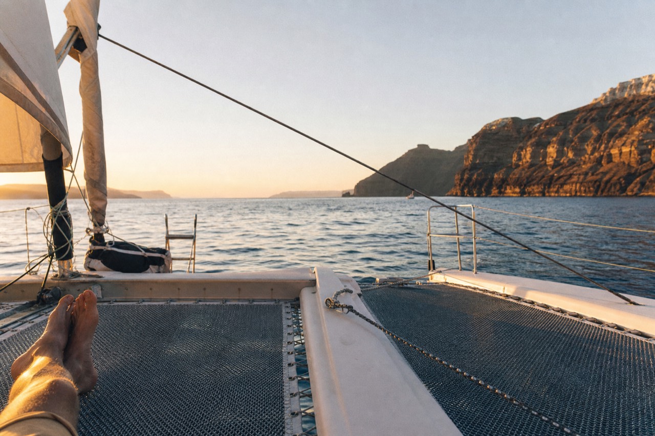 View forward across a catamaran's mesh trampoline nets at golden hour, bare feet and long shadows, turquoise water rushing between the two hulls