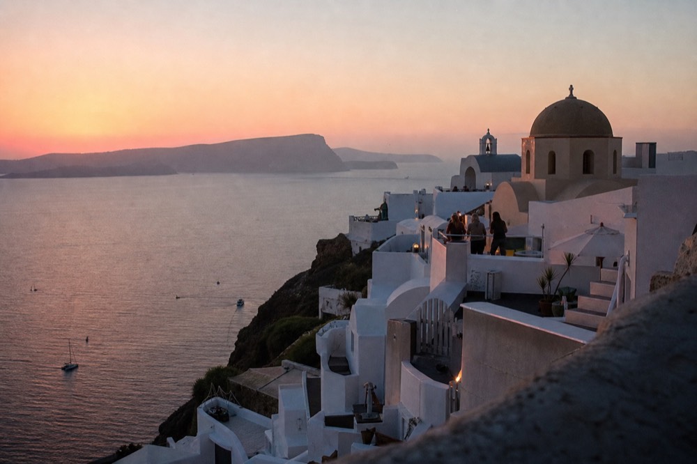 Whitewashed cubic houses and a rounded chapel dome washed rose and gold by low sun, deep caldera water far below the terraces