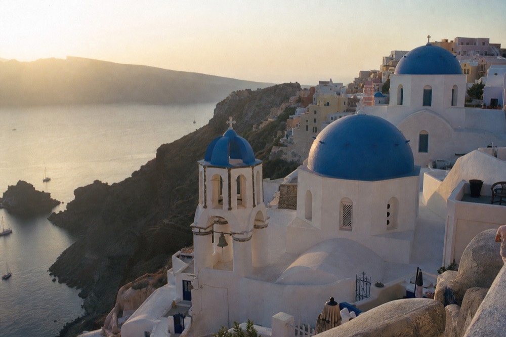 A row of blue-domed chapels above the caldera in early morning light, empty terraces and a hazy sea far below