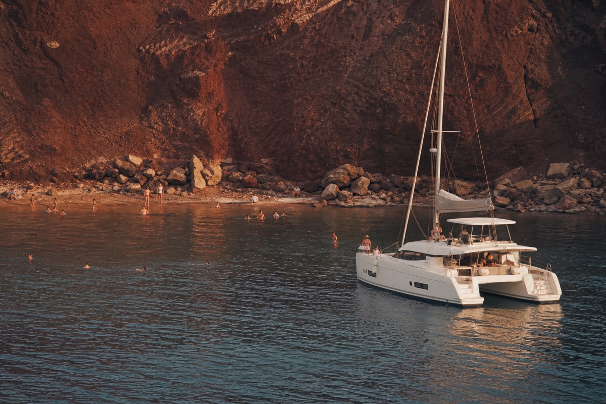 Shared catamaran anchored near the Palea Kameni hot springs in late-afternoon light, swimmers in the rust-tinted water, caldera cliffs rising behind