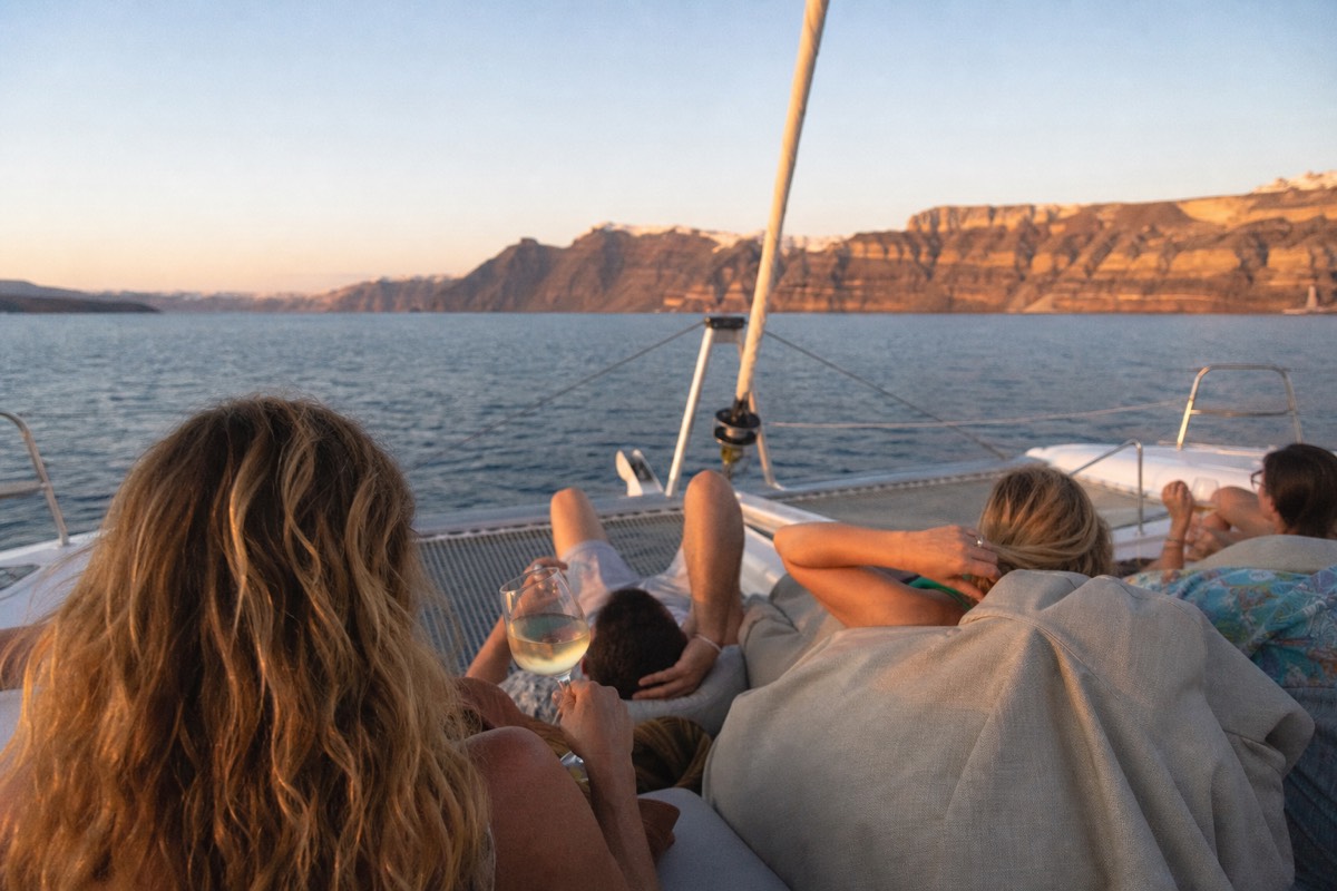 Handful of guests on the bow trampolines of a large sailing catamaran, glasses of white wine in hand, calm Aegean water and evening light