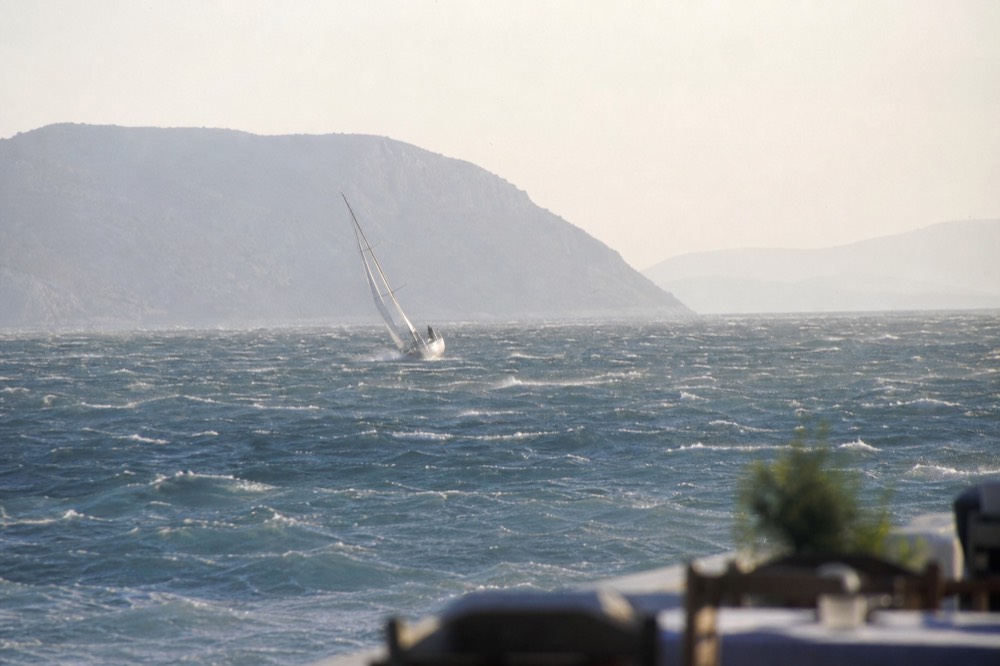 Whitecaps and spray blowing off a hard northerly across open Aegean water, a heeled sailboat small in the distance
