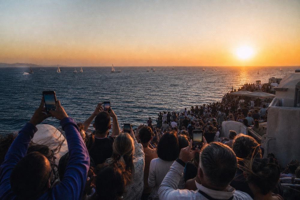 A crowded clifftop terrace packed shoulder to shoulder with people holding phones up toward a setting sun