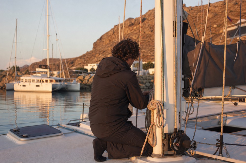 A crew member coiling a halyard at the mast base of a moored catamaran in early morning light, marina water glassy behind