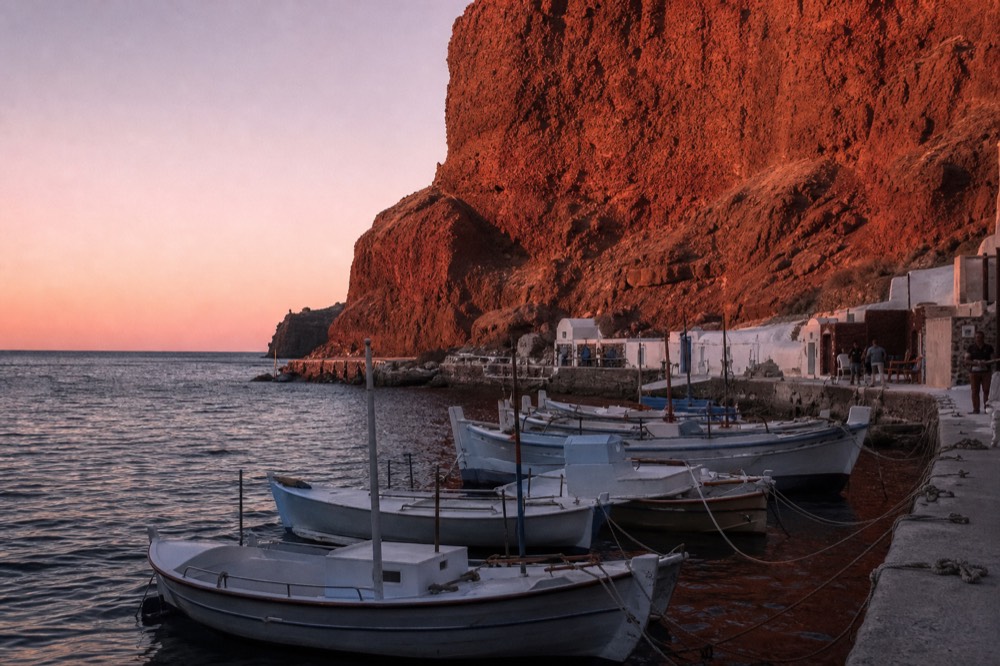 Red iron-stained cliffs towering over a tiny fishing harbour with wooden caiques tied bow-in, late sun turning the rock face deep rust orange
