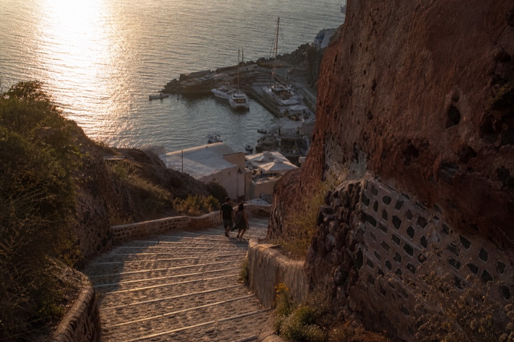 Wide stone switchback steps descending a red cliff face toward a small harbour, two walkers in shadow near the lower bend, sea glittering beyond