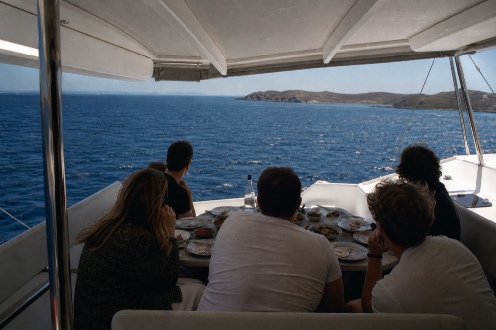 Small group of guests seated around a low table on a catamaran's shaded aft deck, plates of appetizers and a bottle of clear spirit between them, sea beyond
