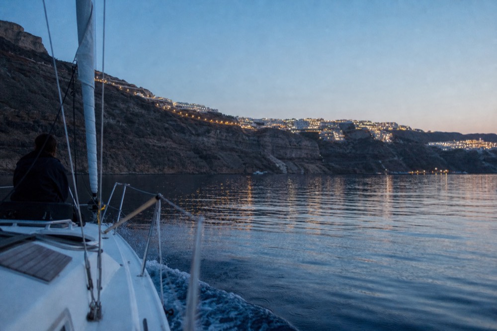Village lights strung along a dark caldera rim during blue hour seen low from a moving boat, faint wake spreading across glassy water