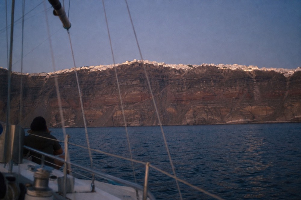 Whitewashed cliff-top houses reduced to a thin bright line along the top of a vast banded lava wall, photographed from sea level in warm evening light