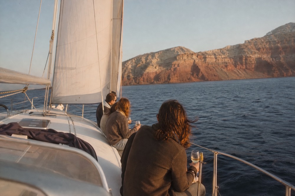 A small group of friends sitting along the windward rail of a sailing yacht, wine glasses in hand, warm side light and open sea behind them