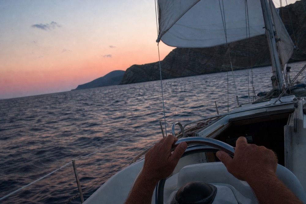 A skipper's hands resting on a stainless steering wheel, horizon tilted slightly, sail edge and a dark cliff wall filling the upper frame