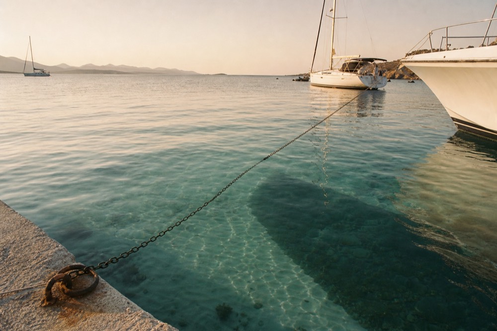 Anchor chain running down into clear turquoise water over a pale sand patch, the dark shadow of a hull printed on the seabed below