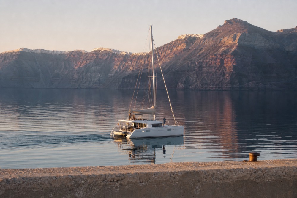 Glassy turquoise water inside the caldera at first light, a single white catamaran motoring past a harbour wall with no wind and a mirrored surface