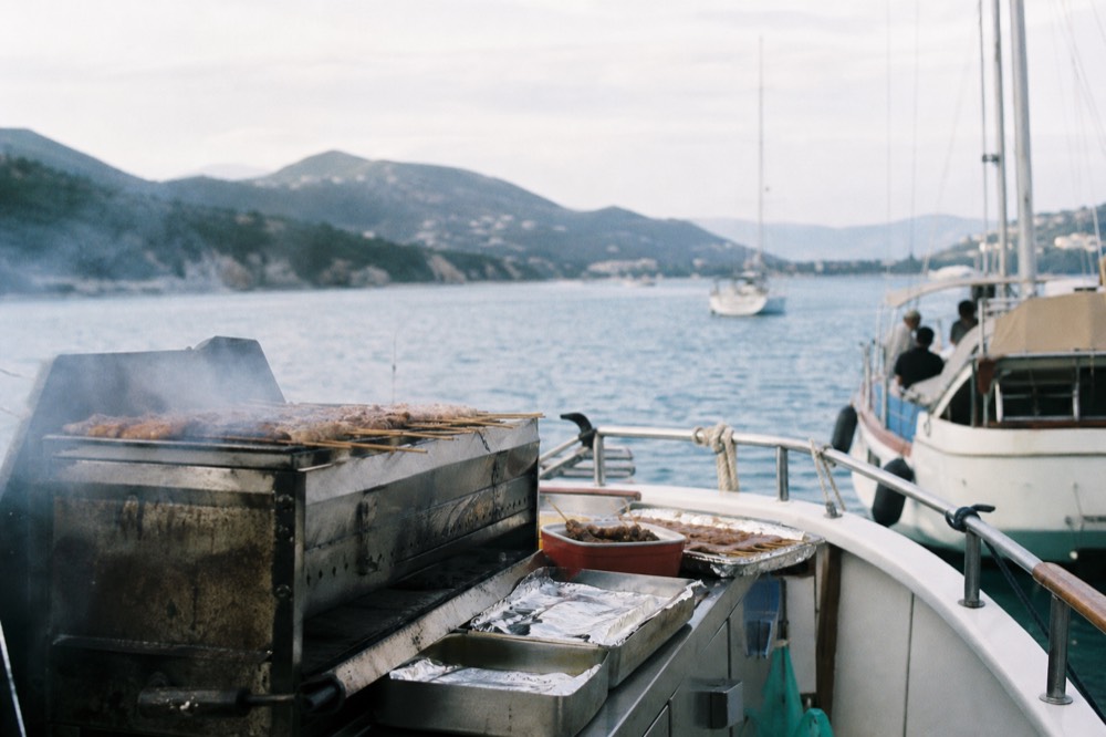 Charcoal smoke lifting off a stern grill on a boat deck, skewers and foil trays laid out on a stainless counter, blurred blue sea behind