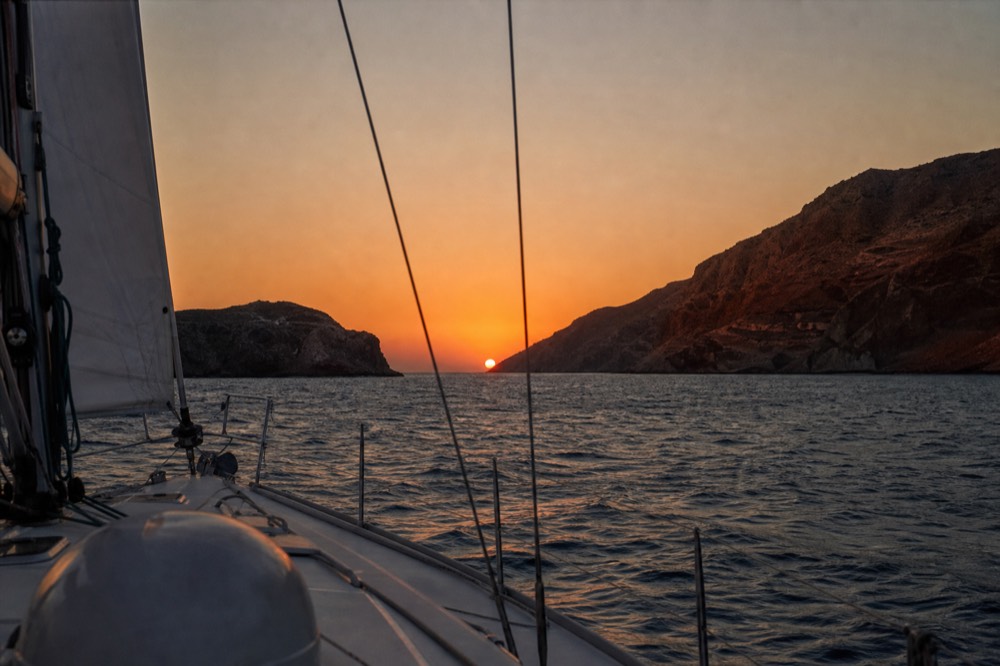 The sun as a flattened orange disc touching the horizon, seen between two mast stays from a moving deck, dark silvered water below