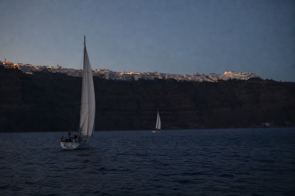 Whitewashed villages seen from sea level as a thin bright ribbon along the top of a dark cliff, deep blue water filling the foreground
