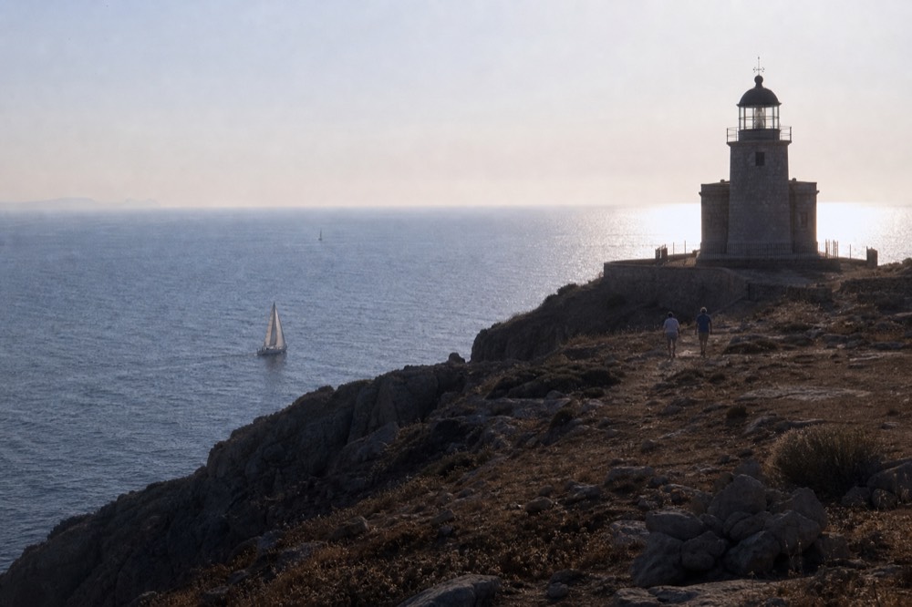 A squat stone lighthouse on a bare headland above dark cliffs, low afternoon sun behind it and deep blue open sea stretching away to the left