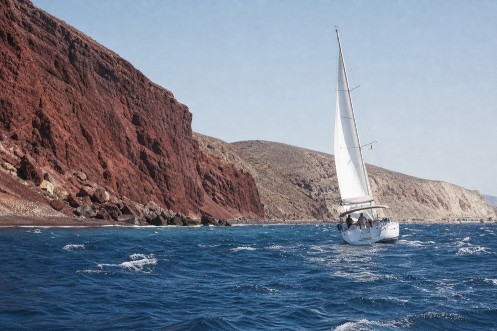 Rust-red cliffs meeting turquoise shallows along a short dark shoreline, seen from offshore with a pale pumice headland further along the coast