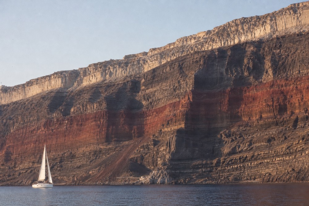 Horizontal bands of black lava, red scoria and pale ash stacked hundreds of metres high above a small white sailboat dwarfed at the base of the cliff