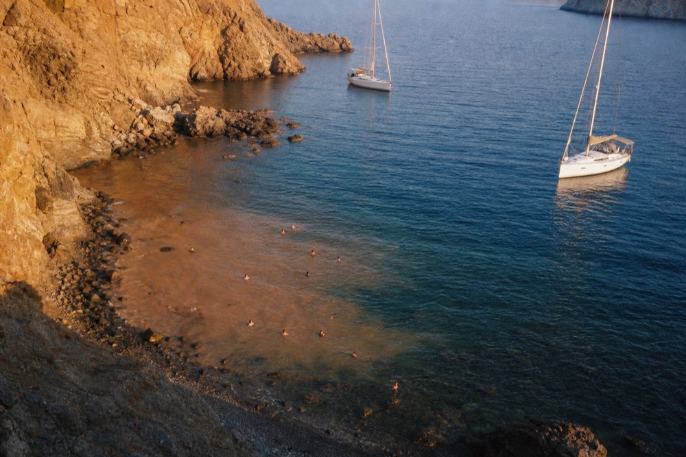 Orange-brown mineral water fanning out from a shallow rocky cove into clear blue sea, ochre cliffs above and swimmers' heads dotted in the warm patch