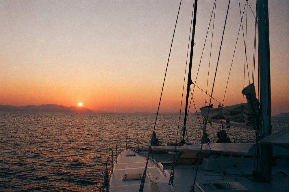 A catamaran's furled sails and rigging silhouetted against a low orange sun over open water, distant island ridge flattening into haze