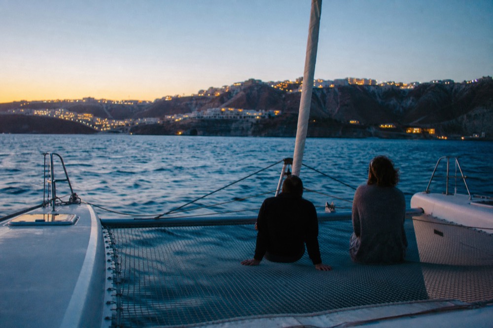 Bare feet and shadows on the taut mesh trampoline between two catamaran bows, deep blue water rushing past underneath in late afternoon light