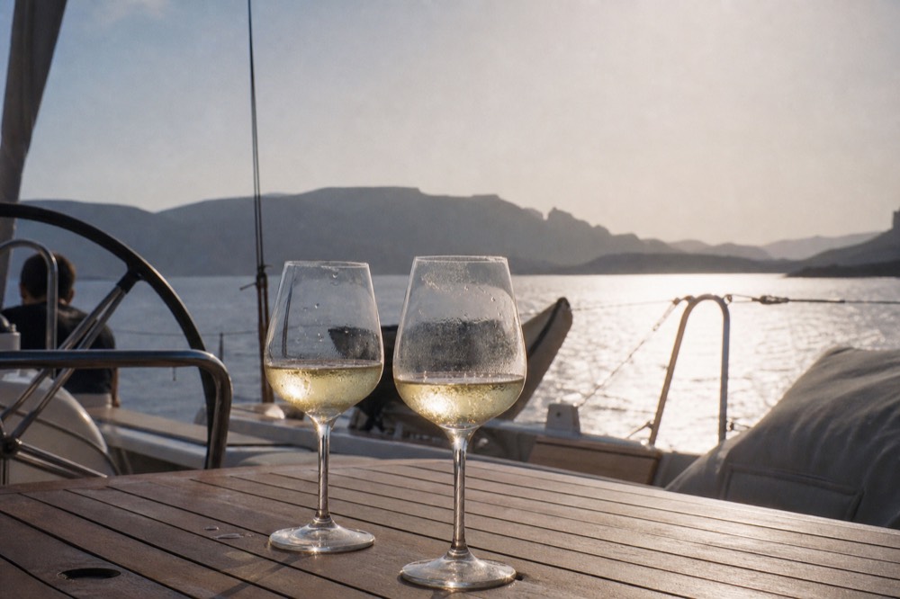 Chilled white wine glasses on a teak cockpit table backlit by low sun, condensation on the rims and the caldera rim blurred behind them