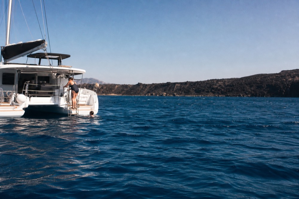Swimmers descending a stainless stern ladder into deep blue water from an anchored white catamaran, ripples spreading toward a dark volcanic shoreline