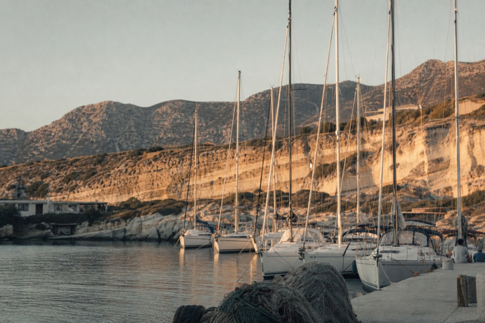 Pale sculpted cliffs and moored white hulls at a small southern marina under hard afternoon light, mast tips lined up against a dusty ridge
