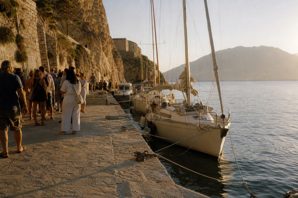 A stone quay crowded with people waiting in the sun beside tied-up tenders, low cliff walls rising straight behind them