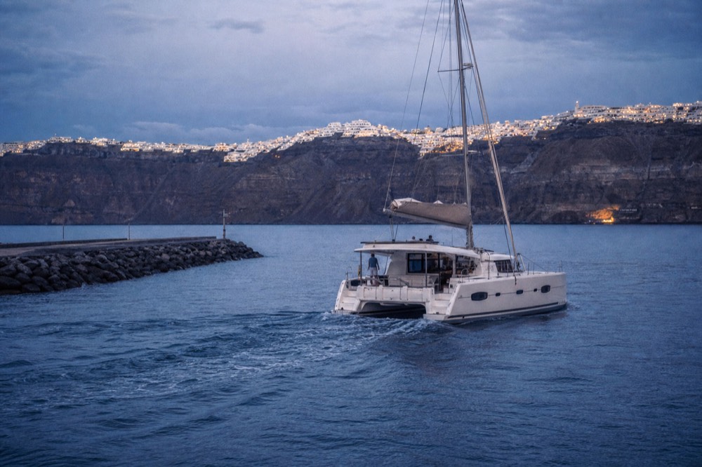 A white catamaran motoring out of a flat southern marina in mid-morning light, breakwater rocks and calm water astern