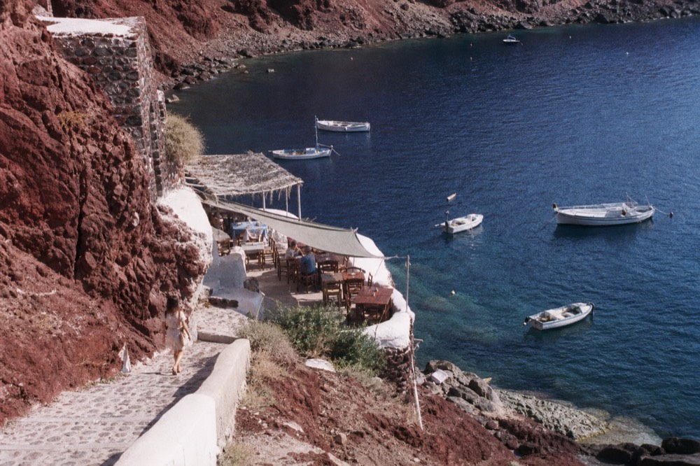 Red cliffs and a stepped path dropping to a tiny bay lined with wooden tables and moored open fishing boats