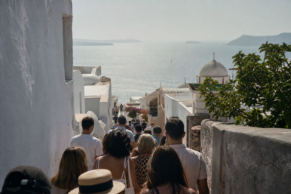 A narrow marble village lane jammed with visitors between white walls at midday, pale domes visible over the rooftops