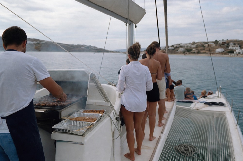 Smoke rising off a stern deck grill on an anchored catamaran, skewers and foil trays on the rail, barefoot guests queueing along the side deck