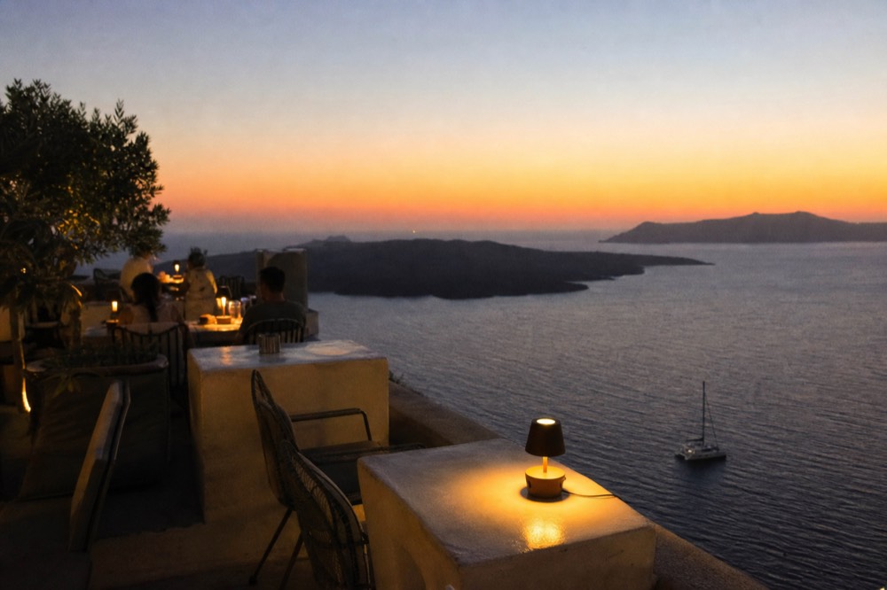 A caldera-view restaurant terrace at dusk, small lamps on stone tables and empty chairs facing the drop, the sea already dark below