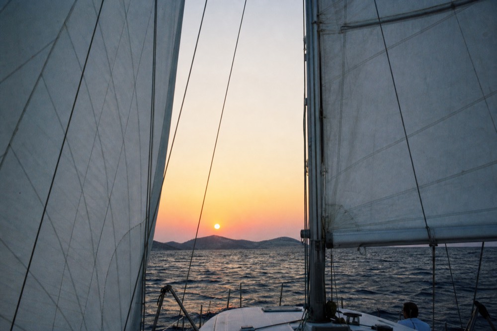 The sun a flattened orange disc just above a low dark island, seen between two taut white sails from a moving deck