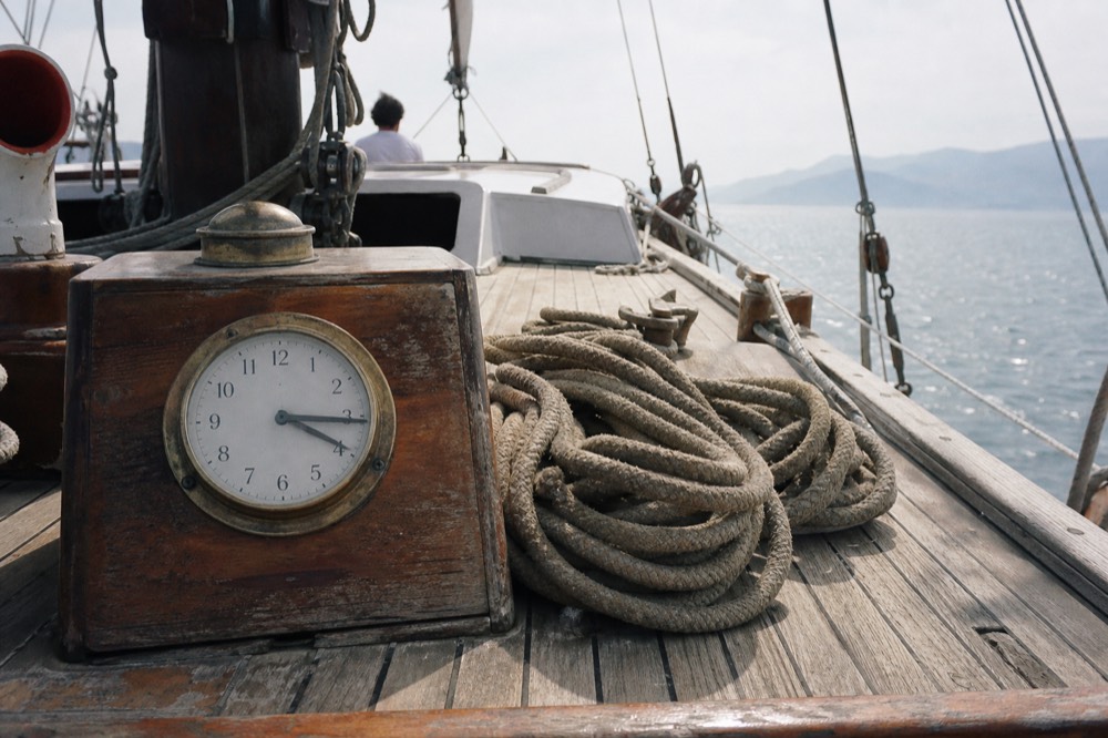 A ship's clock and a coiled mooring line on sun-bleached teak, hard midday shadows across the planking
