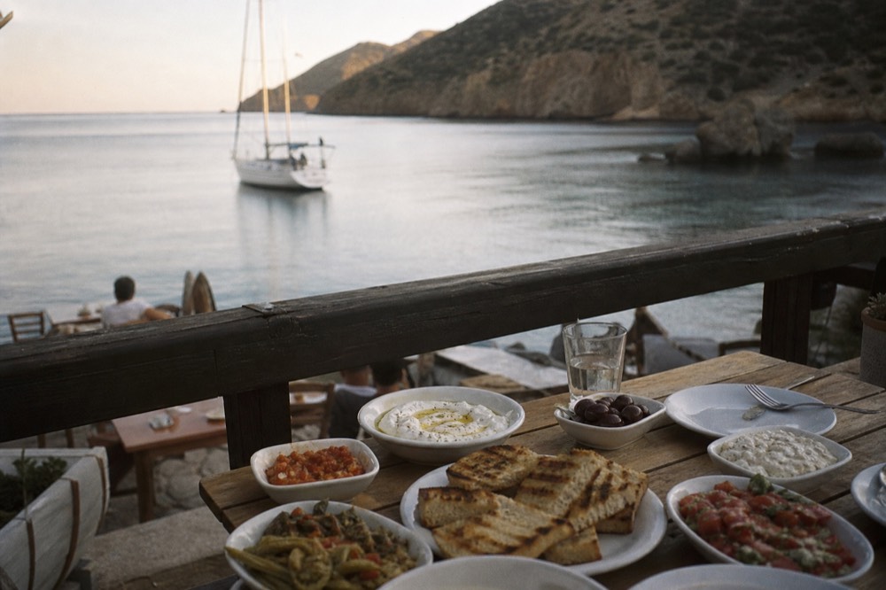 Plates of mezedes, olives, dips and grilled bread crowding a small deck table, sea and dark rock softly out of focus beyond the rail