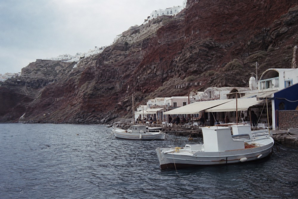 Red volcanic cliffs dropping straight into a narrow bay lined with tavern awnings and moored fishing boats at their base